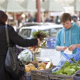 Woman in black coat holding bunch of carrots, stall seller wearing blue hoodie, preparing bag for the produce 