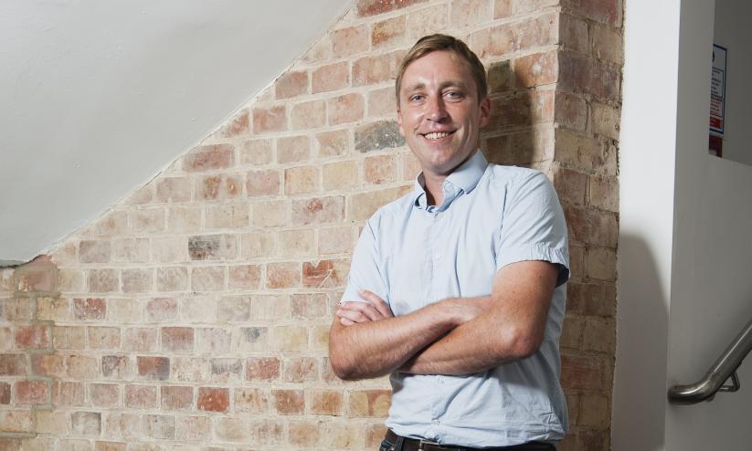 Conrad Peberdy standing infront of brick wall, arms folded, smiling